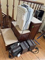 Wooden three-step storage cabinet with three cushioned tops stacked near stair railing, along with three electric space heaters with visible power cords.