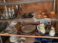 Shelving containing various ceramic tea sets including brown textured set and fruit design sugar bowl; chicken-shaped pitchers; metal cocktail shaker and champagne flutes; plates and chargers; wooden cutting boards.
