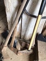 Variety of yard tools leaning against a wooden wall including sledgehammers, pickaxe, axe, hatchet with wooden handles on a concrete floor.