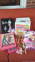 Photo of multiple vintage Barbie doll cases, dolls, clothing, and accessories arranged on an orange sofa. Includes black and white doll cases, dolls without clothes, a sealed pink dress package, and small doll accessories in a bag.