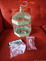Overall view of white hexagonal metal birdcage with two packaged artificial birds on a red upholstered background.