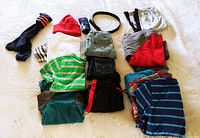 Overview of assortment of boy's clothing including socks, shirts, pants, shorts, and a red knit hat spread on bed