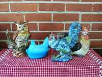 Ceramic rooster wall hanging, ceramic owl statue, blue glass nesting hen, and ceramic rooster statue displayed on red checkered cloth against brick wall.