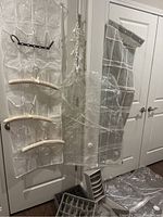 Photo showing two hanging door organizers attached to a white door: one transparent with compartments and black hanger, the other clear with grid compartments. Also visible are cream satin padded hangers and white plastic clothes pin chain hanging.