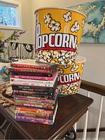 Two large yellow popcorn buckets branded with Bath & Beyond, decorated with popcorn images, stacked on a table next to a collection of DVDs.