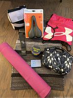 Lot with all items arranged on a floor: bike seat in package, fitness dice, Under Armour drawstring bag, insulated lunch bags, black strap, rolled pink yoga mat