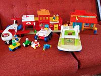 Full view of vintage Fisher Price playsets and accessories arranged on a red couch. Includes airplane, boat, village and Western Town buildings, and multiple peg people with small vehicles.