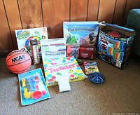 Photo of assorted unopened and opened games and toys on carpet against wooden paneled wall, including basketball, Magic Tracks, Twister, Lego Worlds Switch game, and various boxed games and puzzles.