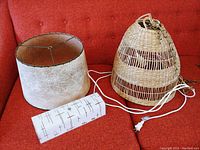 Three lamp shades on red couch: large woven basket hanging shade with chain and cord, Mid Century fiberglass shade with dark stains, and small white rectangular shade with brown design.