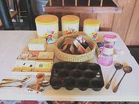 Photo showing three yellow lidded metal canisters with floral decoration, woven basket with small kitchen items, pink and clear plastic grinder and chopper, cast iron muffin tin, and silver plated utensils.