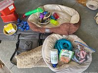 Photo showing two dog beds, one brown/beige Cesar Milan bed with a gold cushion and hole, the other a beige round bed, with multiple colorful dog toys and pet supplies around them.