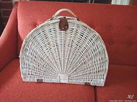 Front view of semi-circular white wicker picnic basket with leather latch and handle, placed on red couch.