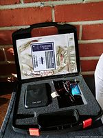 Open black foam case showing TENS 3000 device, electrode pads packet, battery, and wires.