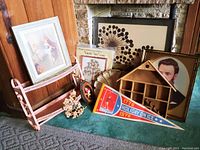 Various wall hangings and decorative items arranged on carpet against a fireplace and wood panel wall, showing signed Alan Buckle dandelion art, family tree kit, soldier portrait, wooden shadowbox, chalkware shell, and pennant.