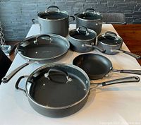 Photo showing assortment of Calphalon saucepans, sauté pan, frypans, stockpot on a white table with glass lids and metal handles.