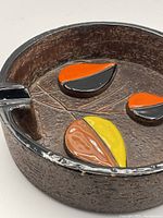 Close-up showing the textured brown surface, black rim, and glossy colorful ceramic leaf shapes in orange, black, yellow, and brown.