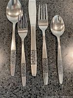 Close up photo showing dinner knife, dinner fork, dessert fork, soup spoon, and teaspoon with detailed scrolled floral engraved handles.