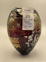 Front view of the burgundy glass vase showing gold, white, and iridescent details and the label indicating it is signed by Tim Lazer and about 10 inches tall.