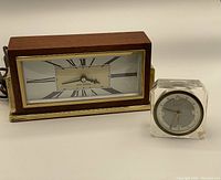 Two clocks side by side: mid-century modern Seth Thomas electric clock with wood and brass, and small acrylic Elg-Art clock.