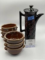 Photo shows entire lot including tall dark pottery coffee pot with circular patterned texture and 6 stacked brown rooster motif bowls with handles.