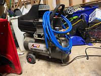 Full view of American Force Pro 2.5 HP air compressor with blue hose coiled around handle, wheels visible, showing signs of  use and dust.
