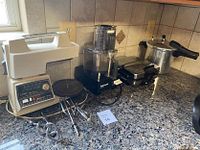 Four assorted kitchen appliances on granite countertop, showing Oster stand mixer, Cuisinart food processor, Black & Decker waffle iron, and pressure cooker.