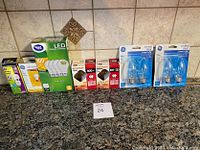 Assorted unopened lightbulbs with original packaging arranged on countertop, showing various brands including GE, Great Value, and Feit Electric. Bulbs include LED, incandescent, and decorative flame tip types.