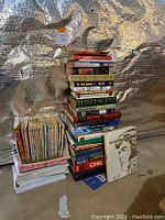 Two stacks of assorted used books and several unopened packs of lined paper in plastic wrap on floor against a silver reflective background.