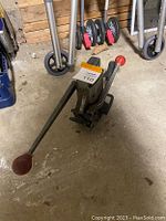 Black hydraulic floor car jack with red handles on the lifting arm and long handle for pumping, placed on a concrete floor in a workshop environment.