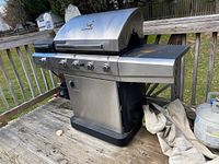 Char-Broil gas grill with four burners and side burner on left, stainless steel finish, outdoor condition with some wear, closed lid and bottom storage compartment.
