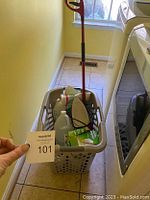 Photo of the filled grey laundry basket with cleaning supplies and mop handle visible, placed on tiled laundry room floor.