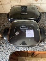 Two electric skillets placed on a countertop, one Farberware and one WestBend, both with glass lids and side handles.