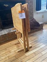Side view of two folded wooden tray tables standing upright in their matching wooden stand on a hardwood floor.