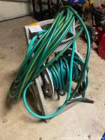 Two green garden hoses, one green and yellow stripped hose loosely coiled and the other plain green hose wound around a grey plastic holder. The items are placed on a floor with other items visible in the background.