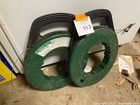 Two partially used Greenlee steel fish tapes on green reels with handles, resting against a white wall on a garage floor.