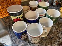 Photo of 9 holiday themed mugs clustered together, showing various festive patterns including poinsettia, holly, and snowmen on ceramic mugs in assorted colors.