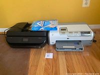 Pair of HP printers side by side on wooden floor, with a pack of paper on top