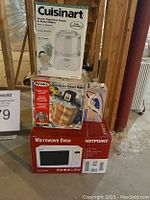 Photo showing boxed Cuisinart Frozen Yogurt-Ice Cream & Sorbet Maker, Rival Electric Ice Cream Maker, Hotpoint Microwave Oven, and a boxed iron stacked on the floor in a warehouse-like setting.