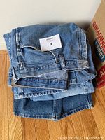 Stack of five pairs of men's jeans folded on a hardwood floor, showing multiple denim shades from light to dark blue. One pair has a size tag visible.