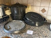 Photo showing black speckled metal Dutch oven with lid, steamer pot set with stackable components, and a round metal griddle on a kitchen counter.