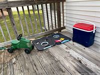 Shows the outdoor toy bundle including the green kid’s riding tractor, black corn hole boards with bean bags, and the blue and red Igloo cooler on a wooden deck
