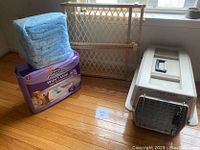 Pet bundle showing Petmate Pet Taxi Carrier, wooden pet gate, and a large pack of potty training pads on wooden floor near window.