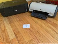 Both printers photographed on a wooden floor, Canon on the left and HP Deskjet D1420 on the right, showing overall condition and sizes