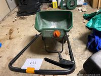 Front view of green Scott's Standard Broadcast Spreader showing hopper, orange adjustment knob, part of black metal frame and wheels.