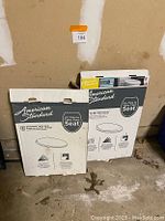 Photo of two American Standard toilet seat sets in packaging standing on concrete floor against wall.