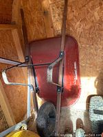 True Temper USA wheelbarrow leaning against wooden wall showing red metal tub, wooden handles, metal frame, and rubber tire.