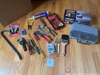 Photo showing various household tools including Dremel case, angle grinder box, paint brushes, screwdrivers, wrenches, crowbar, drill bits, and other hand tools on wooden floor.