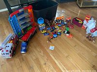 Overview of various toy vehicles on a wooden floor including fire truck, school bus, monster trucks, and other assorted cars. Black container and plastic jar with blue lid also visible.
