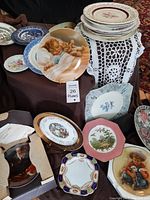 Overview of assorted decorative plates arranged on a table and stack, showing multiple artistic themes and styles including cherubs, children, florals