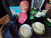 Photo showing close-up of some teacups, doll in pink crocheted outfit, green and blue decorated ceramic pitcher, vintage sewing machine figurine, and Bamm Bamm book